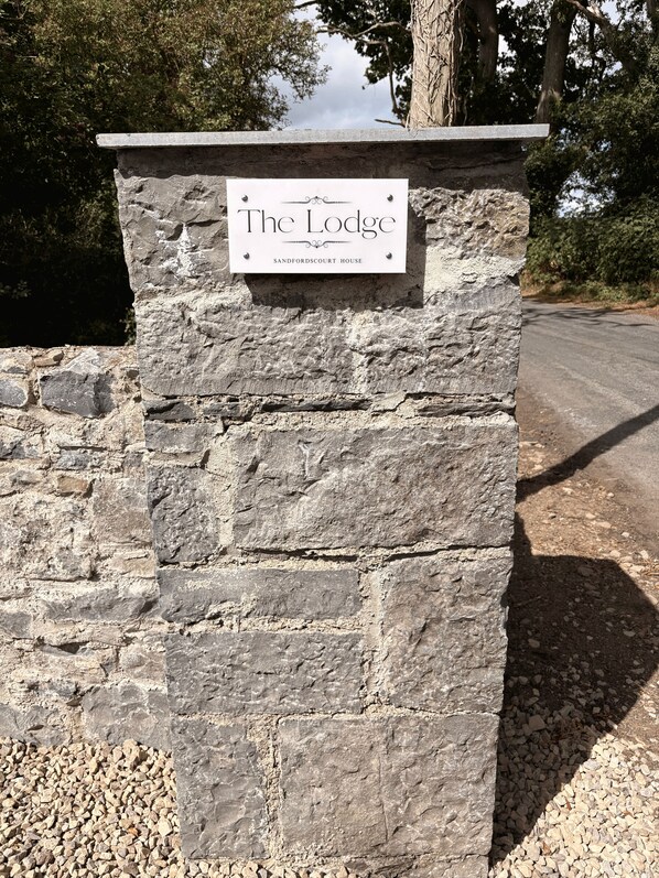 A stone sign with the text 'The Lodge Sandfordscourt House' mounted on a stone wall.