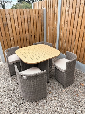 Une table en bois, entourée de quatre chaises en osier, posée sur un sol de gravier et délimitée par une clôture en bois.