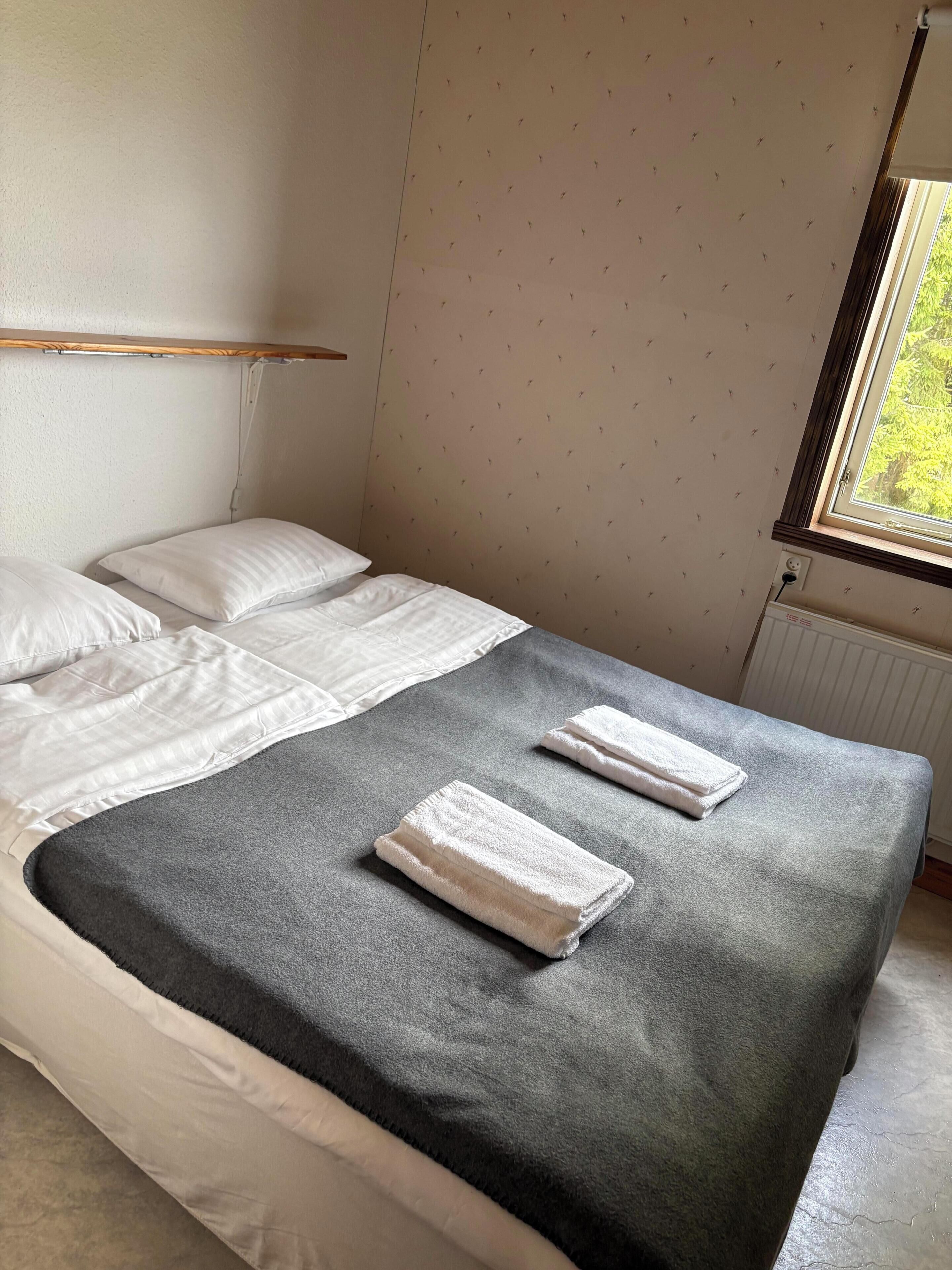 A single bed with white linens and a grey blanket, a window with a view of greenery, and a wall with a subtle pattern.