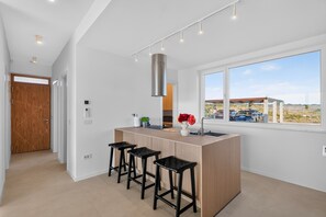 A modern kitchen with a central island, bar stools, a large window, and a view of a desert landscape. - Villa Salis with heated pool & sea view (Košljun)