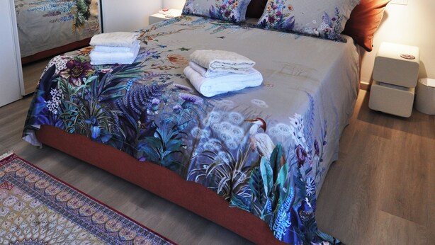 Quarto com cama, travesseiros, mesa de cabeceira, luminária na parede e espelho.