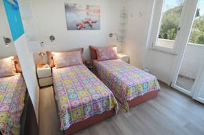 A bedroom with two single beds, a nightstand with a lamp, a wall-mounted shelf, and a window with a view. - Juliet's House (Cagliari)