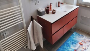 Um banheiro moderno com um porta-toalhas branco, um espelho sobre uma pia branca com torneira de cobre, e piso de madeira.