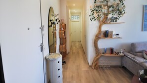Um corredor com piso de madeira, uma porta branca, um espelho e uma estante em formato de árvore com livros e objetos decorativos.