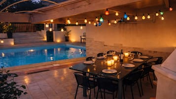 Un espace extérieur aménagé pour les repas, avec une piscine, des guirlandes lumineuses et une table dressée pour un repas.