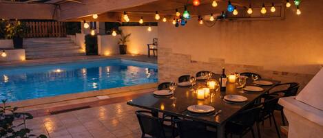 Área de refeições ao ar livre com piscina, luzes de corda e mesa posta para uma refeição.