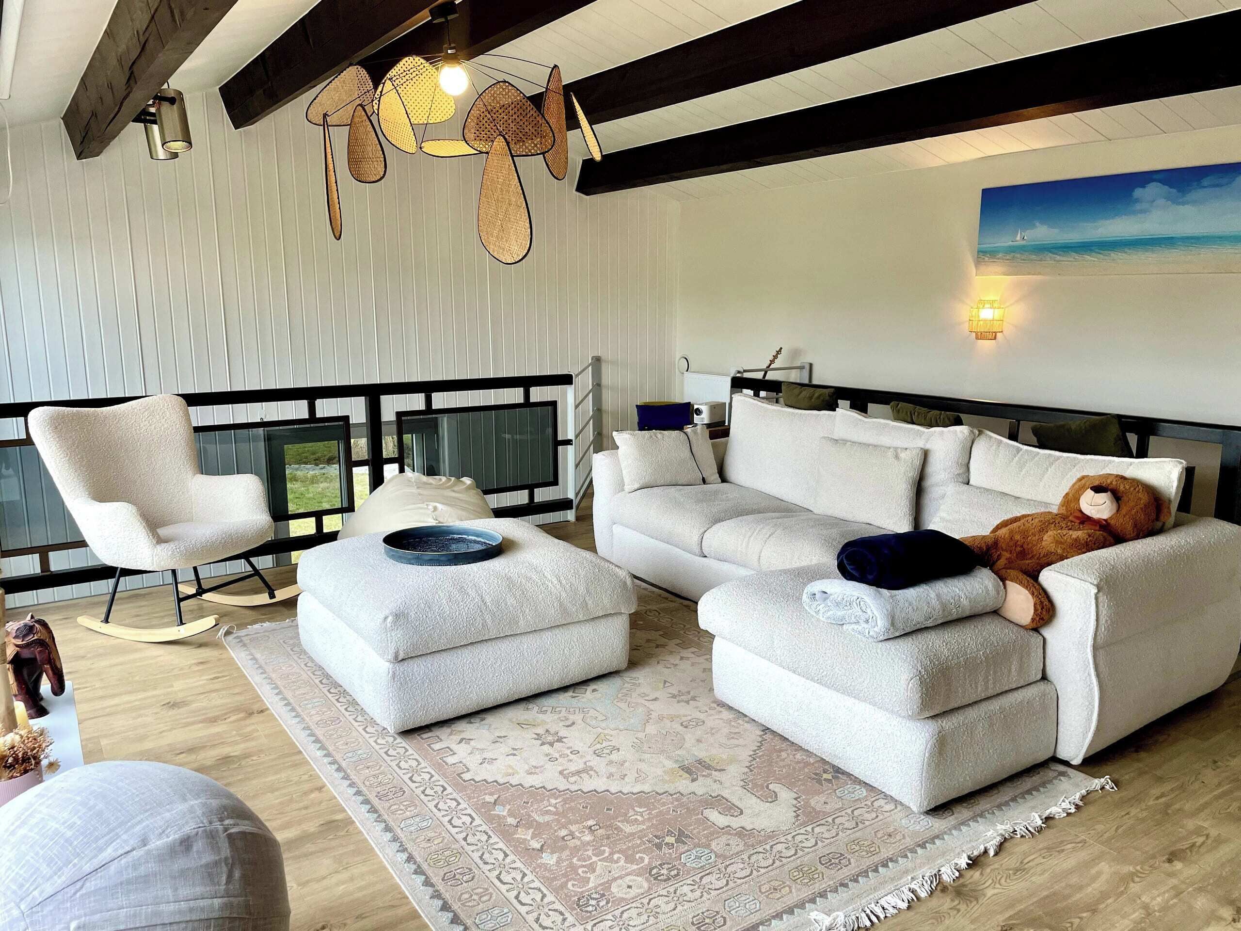 Una sala de estar moderna con sofá, sillón y reposapiés, un tapete decorativo y un cuadro de una playa.