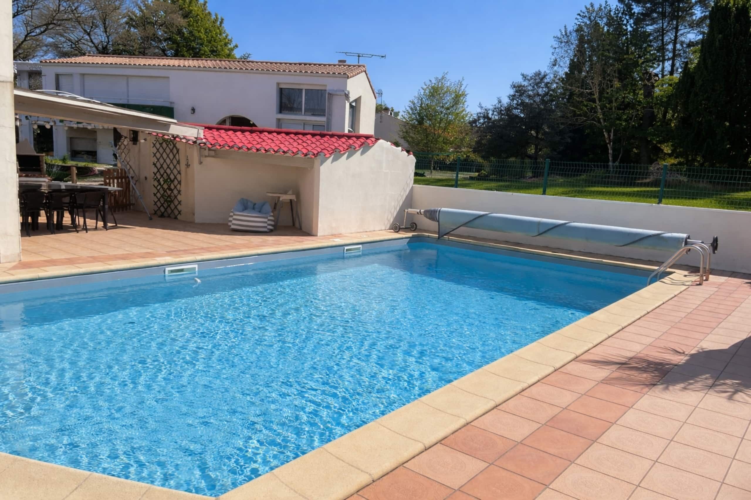 Une piscine avec une terrasse carrelée, un espace salon couvert et une maison en arrière-plan.