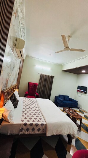 A hotel room with a bed, a red armchair, a blue sofa, a ceiling fan, and a flat-screen TV mounted on the wall. - Kings pride Aravali (Udaipur)