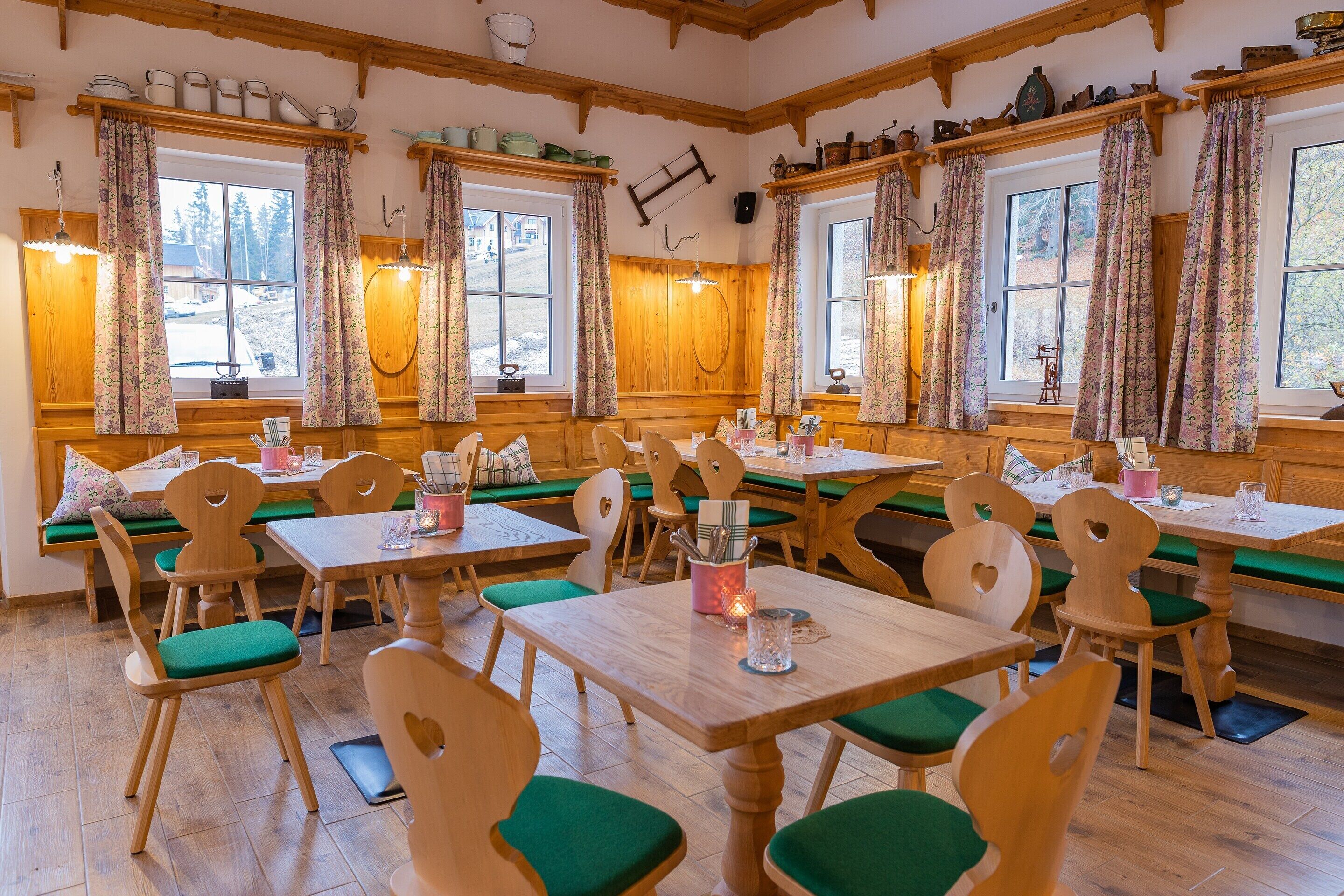 Ein gemütlicher Essbereich mit Holztischen und -stühlen, grünen gepolsterten Bänken und großen Fenstern mit floralen Vorhängen.