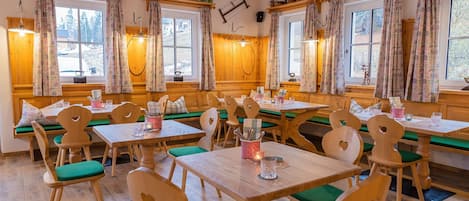 A cozy dining area with wooden tables and chairs, green cushioned benches, and large windows with floral curtains.