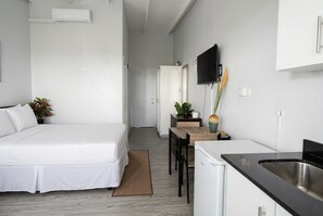 Ein kleines Hotelzimmer mit einem Bett, einem Esstisch, einer Küchenzeile inklusive Spüle und einem an der Wand befestigten Fernseher.