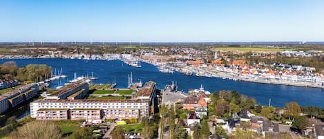 Un pueblo costero con un puerto deportivo, edificios residenciales y aerogeneradores al fondo.