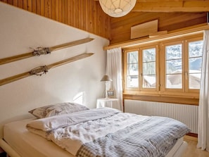Quarto com teto de madeira, cama com roupa de cama branca, janela com cortinas e radiador.