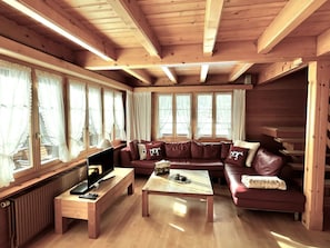 A cozy cabin interior with a wooden ceiling, large windows with curtains, a leather sofa, a coffee table, and a television. - Chalet Rotebächli Appartement/Fewo, Bad, WC, 2 Schlafräume 4-Bettwohnung by Interhome (Lenk)