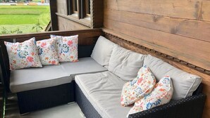 A wicker sofa with white cushions and floral-patterned pillows, positioned on a porch with a view of a green field. - Chalet Rotebächli Appartement/Fewo, Bad, WC, 2 Schlafräume 4-Bettwohnung by Interhome (Lenk)
