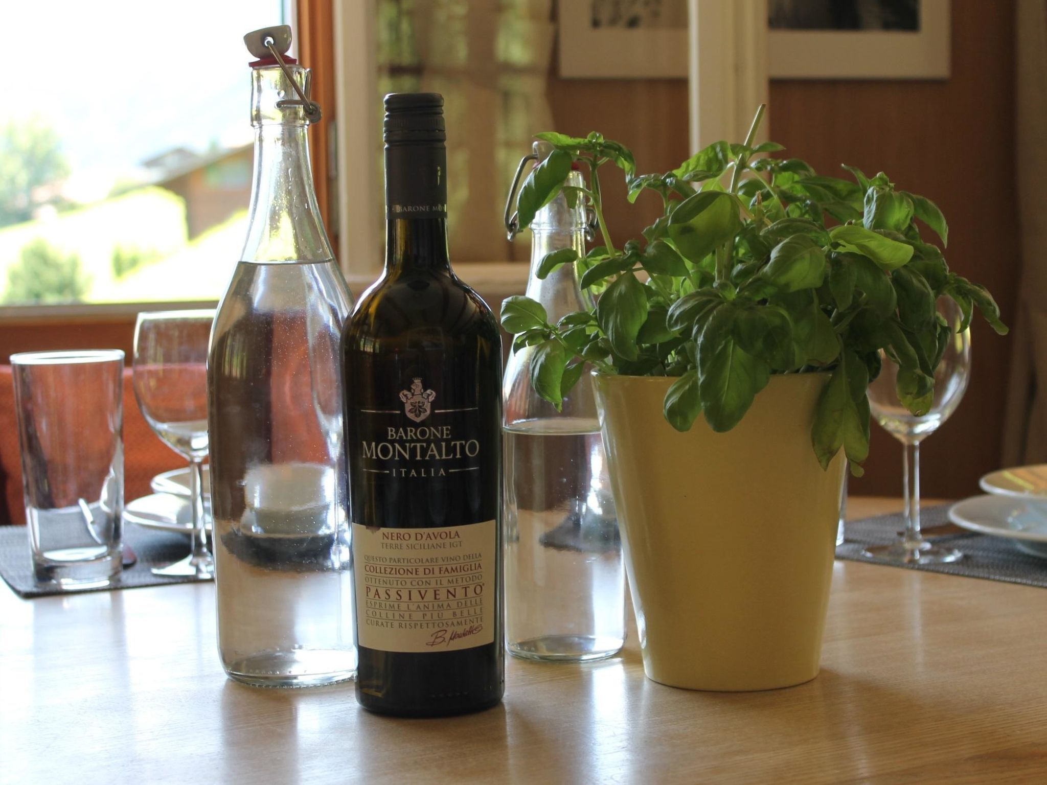 Uma mesa com uma garrafa de vinho Barone Montalto, um frasco de vidro, um vaso de planta e copos de vinho vazios.