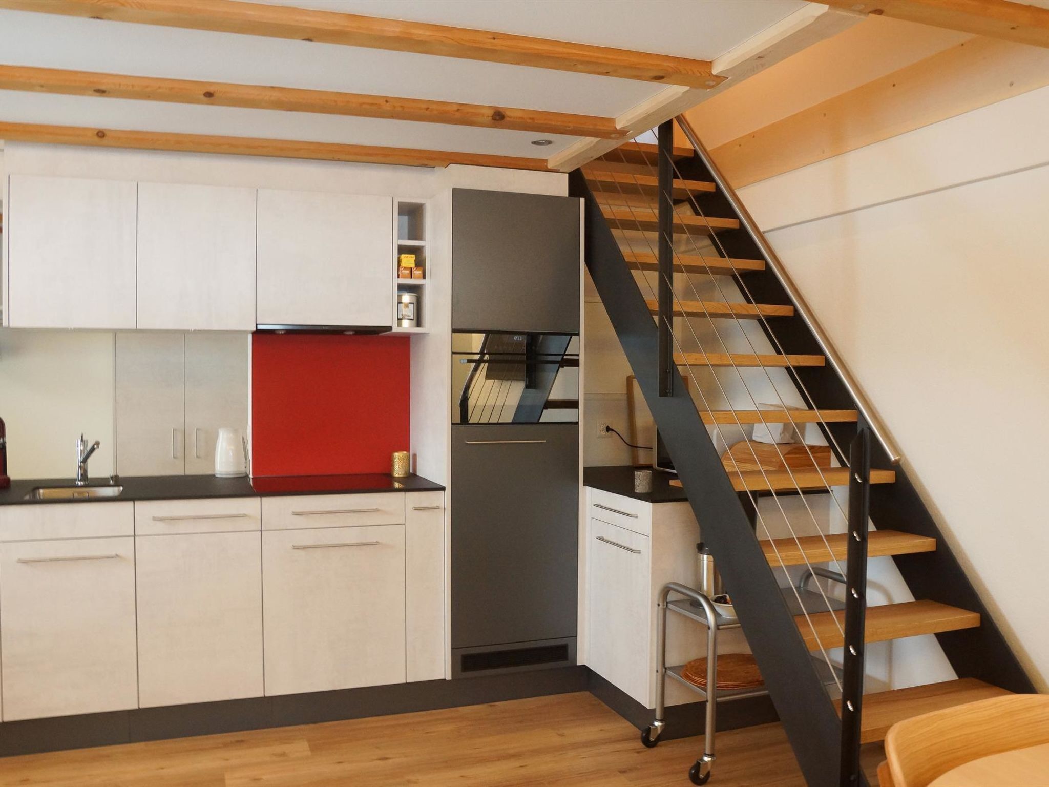 Une cuisine moderne avec des meubles blancs, un mur d’accent rouge et un escalier noir.
