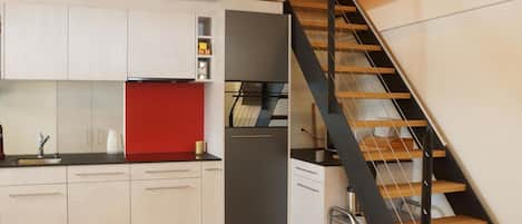 A modern kitchen with white cabinets, a red accent wall, and a black staircase.