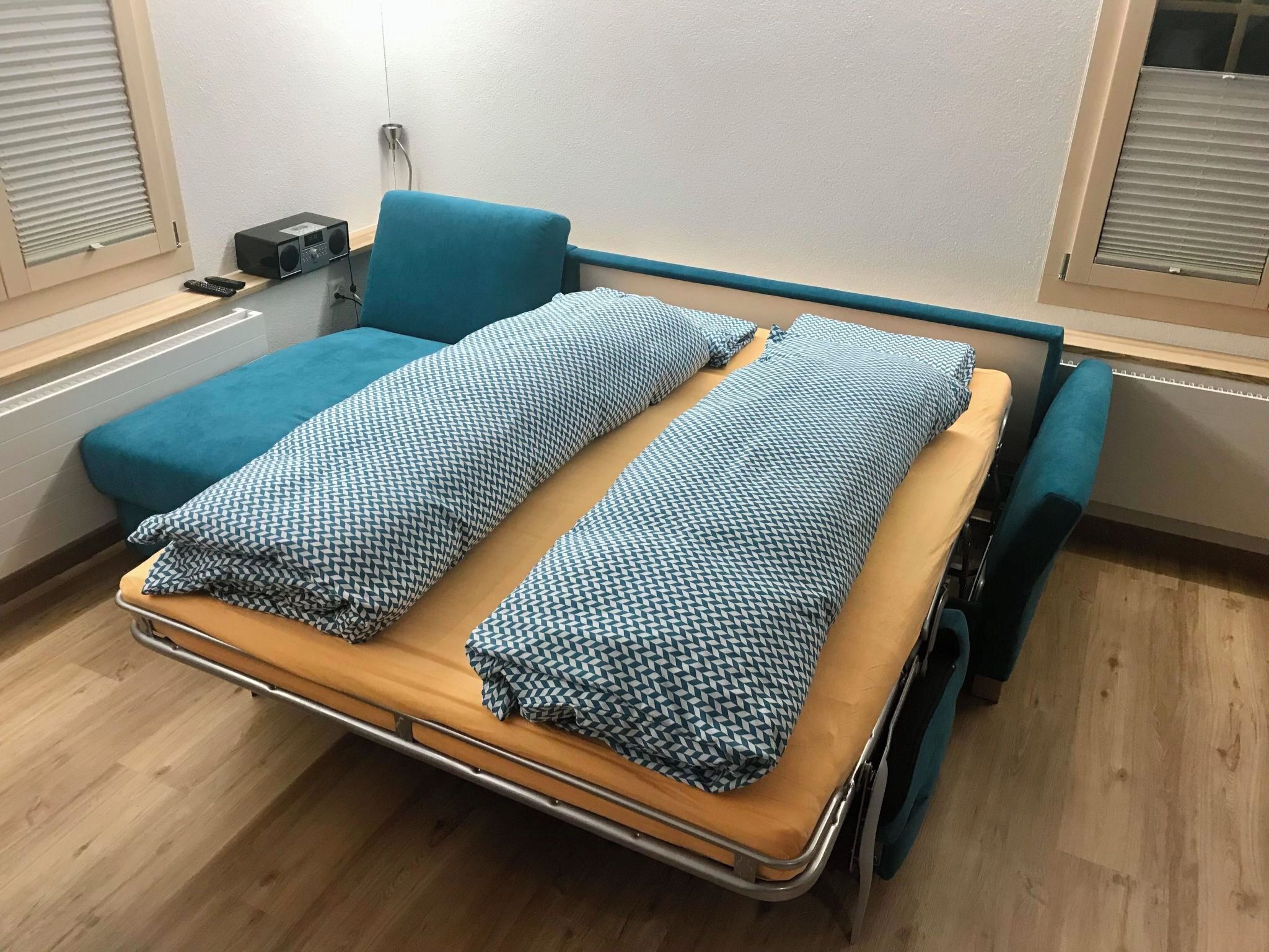 A hospital room with a bed, a small bedside table, a radio, and a window with blinds.