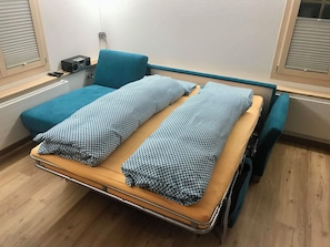 Quarto de hospital com cama, uma mesinha de cabeceira, um rádio e uma janela com persianas.
