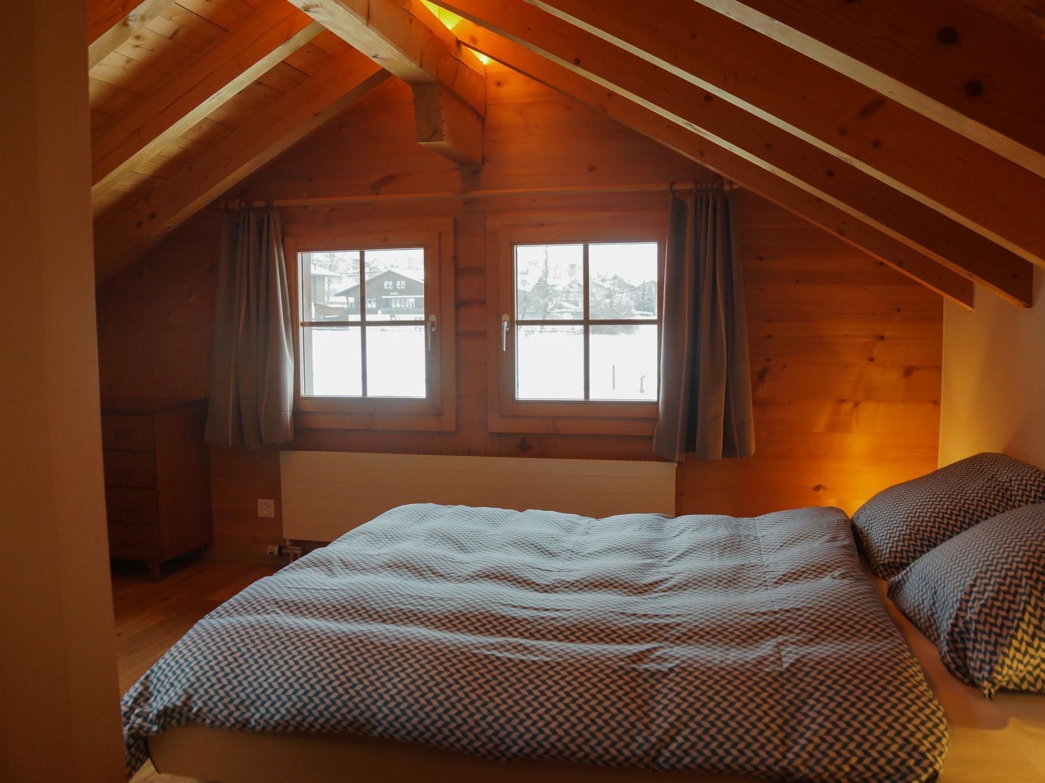 A cozy attic bedroom with a sloped ceiling, a large bed with patterned bedding, two windows with curtains, and a view of a snowy landscape.
