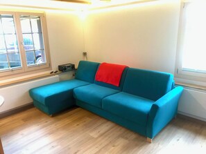 A teal sofa with a red throw blanket, a radio, and a window with blinds. - Lischenhaus 2 - Wohnung Flösch Appartement/Fewo, Dusche, WC, 1 Schlafraum by Interhome (Lenk)