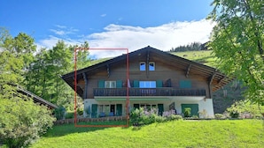Eine zweistöckige Chalet mit Balkon, umgeben von üppiger Vegetation und blauem Himmel.