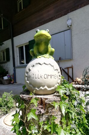 A frog statue on a spherical stone with 'WILLK OMMEN' inscribed, placed on a wooden stump surrounded by greenery. - Chalet Flöschhorn Appartement/Fewo, Bad, WC, 3 Schlafräume by Interhome (Lenk)