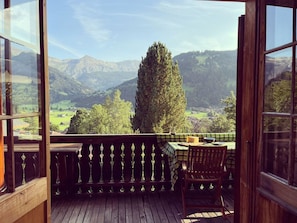 Eine Holzterrasse mit Tisch und Stühlen, mit Blick auf eine gebirgige Landschaft, einen großen Baum und ein Dorf darunter.