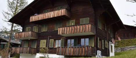 A multi-story wooden chalet with balconies, set in a grassy area with a mountain backdrop.
