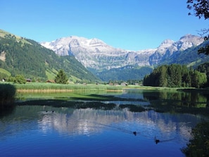 Um lago tranquilo com montanhas ao fundo, rodeado por vegetação e alguns edifícios.