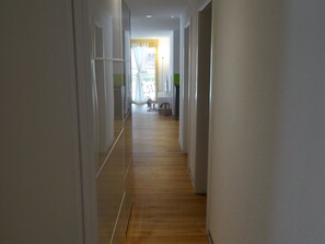 Ein schmaler Korridor mit Holzboden und weißen Wänden, der zu einem Raum mit Fenster und grüner Wand führt.