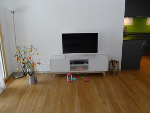 Una sala de estar con un televisor de pantalla plana sobre un soporte blanco, una planta en maceta y una pequeña zona de juegos con juguetes.