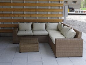 Ein modulares Sofa mit passendem Hocker und einem Korbstuhl, vor einer Betonwand mit horizontalen Holzdielen.