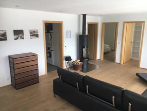 Ein modernes Wohnzimmer mit einer schwarzen Couch, Holzböden, einem Kamin und einem durch eine offene Tür einsehbaren Küchenbereich.