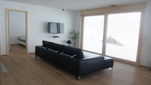 A modern living room with a black sofa, a flat-screen TV, and a large window with blinds. - Chalet Coco Appartement/Fewo, Dusche, WC, 3 Schlafräume 6-Bett-Wohnung by Interhome (Lenk)