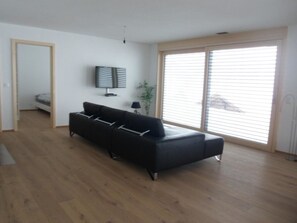 Ein modernes Wohnzimmer mit einem schwarzen Sofa, einem Flachbildfernseher und einem großen Fenster mit Jalousien.