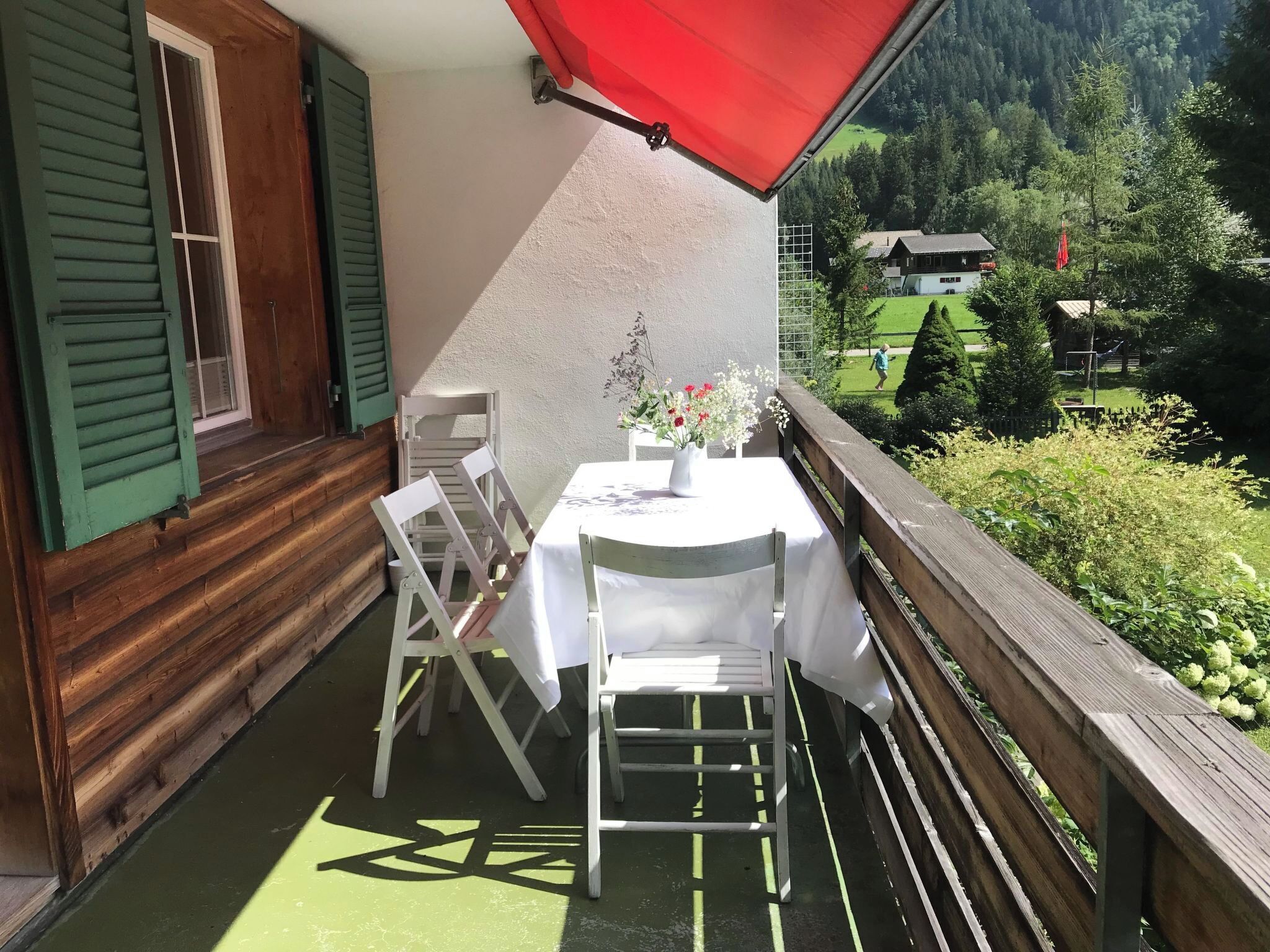 Un balcó amb taula i cadires, una tenda vermella i vista a un paisatge verd amb cases.