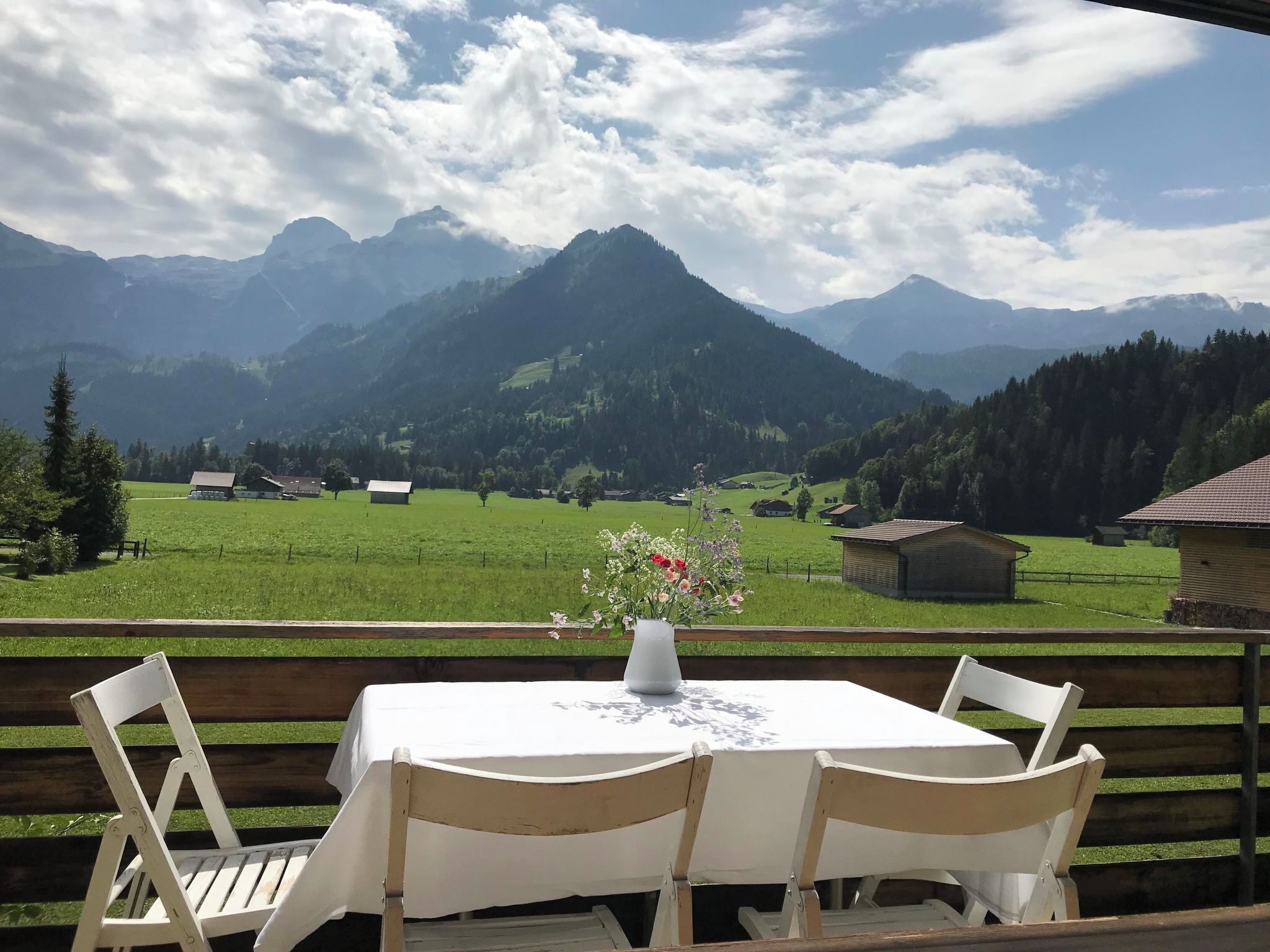 A table with a vase of flowers, two chairs, and a view of mountains and a green field.