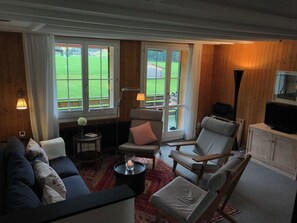 A cozy living room with a sofa, armchairs, a small table with a candle, and a television on a cabinet. Large windows offer a view of a green landscape. - Burgbüel # 1 Appartement/Fewo, Dusche oder Bad, WC, 4 oder mehr Schlafräume by Interhome (Lenk)