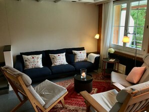A cozy living room with a dark blue sofa, white cushions, a wooden chair, a small round table with a candle, and a view of greenery through a window. - Burgbüel # 1 Appartement/Fewo, Dusche oder Bad, WC, 4 oder mehr Schlafräume by Interhome (Lenk)