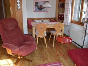 A room with a red chair, a small table, a wooden dining table with chairs, a radiator, and a window with curtains. - Elena # 1 Appartement/Fewo, Dusche oder Bad, WC, 1 Schlafraum 2-Bett-Wohnung by Interhome (Lenk)