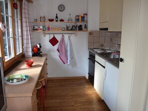 Eine kompakte Küche mit Holzboden, integriertem Backofen und Spüle. Es gibt Hocker an der Arbeitsplatte und ein Fenster mit Vorhängen.