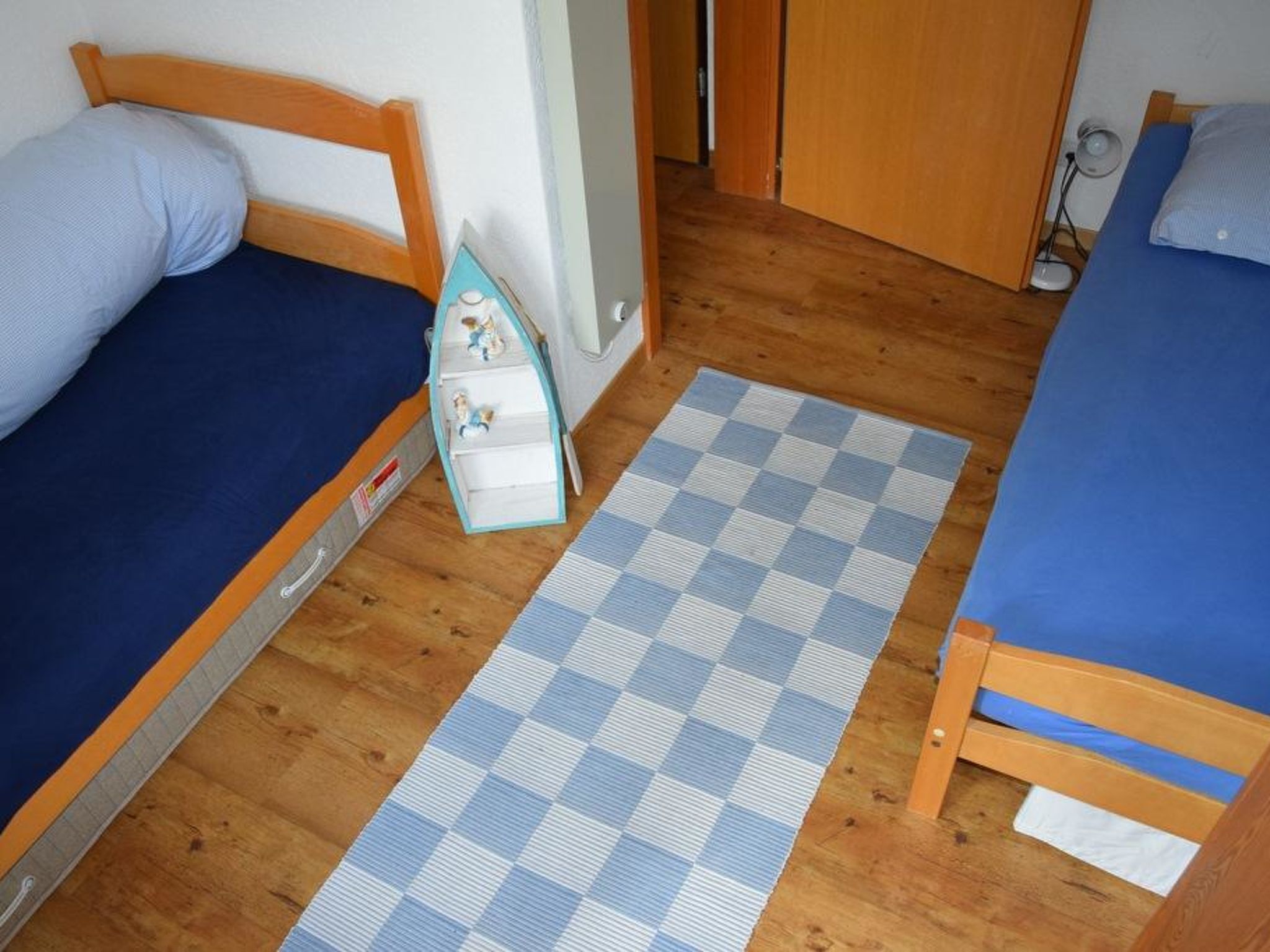 Une chambre avec deux lits simples, un plancher en bois, un tapis à carreaux et une petite armoire blanche.