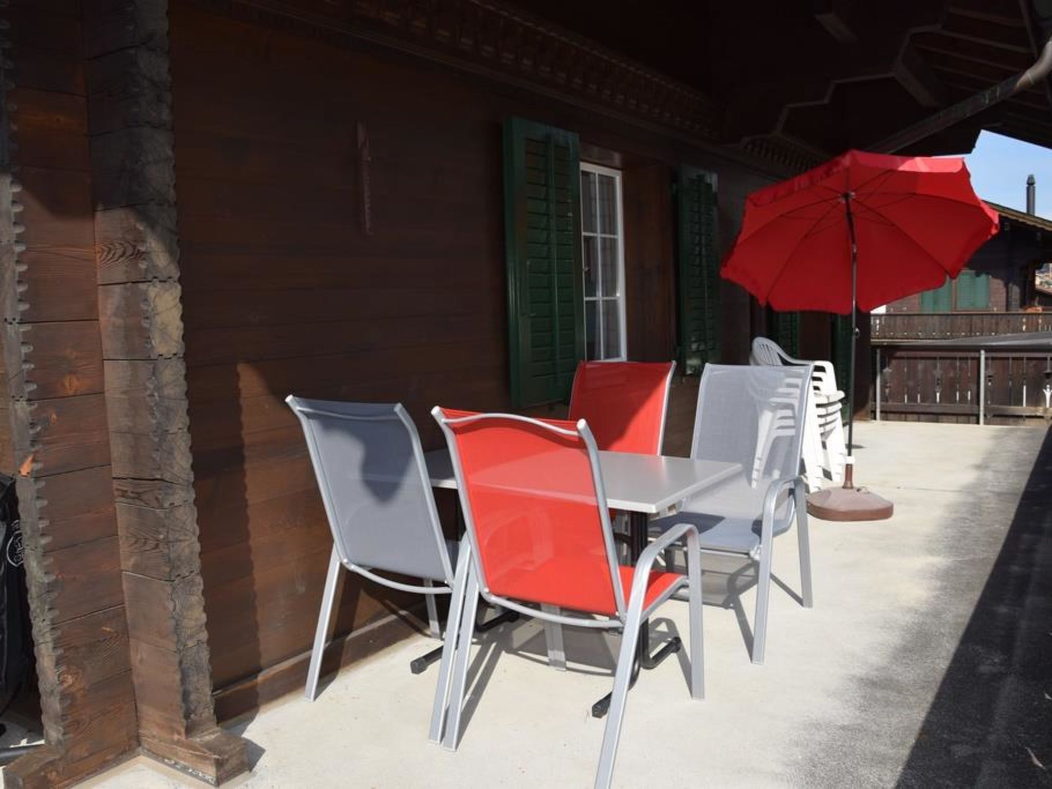 Une terrasse avec une table et des chaises, un parasol rouge et un bâtiment en bois avec des volets verts.