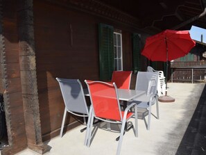 Eine Terrasse mit Tisch und Stühlen, einem roten Sonnenschirm und einem Holzhaus mit grünen Fensterläden.