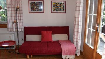 Une pièce chaleureuse avec un canapé rouge, deux œuvres d’art encadrées accrochées au mur, une petite table rouge et une fenêtre avec des rideaux.