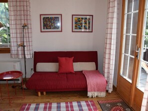 Ein gemütliches Zimmer mit einem roten Sofa, zwei gerahmten Kunstwerken an der Wand, einem kleinen roten Tisch und einem Fenster mit Vorhängen.