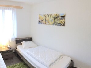 Uma cama de solteiro com colcha branca, uma mesa de cabeceira com abajur e um quadro de árvores na parede.